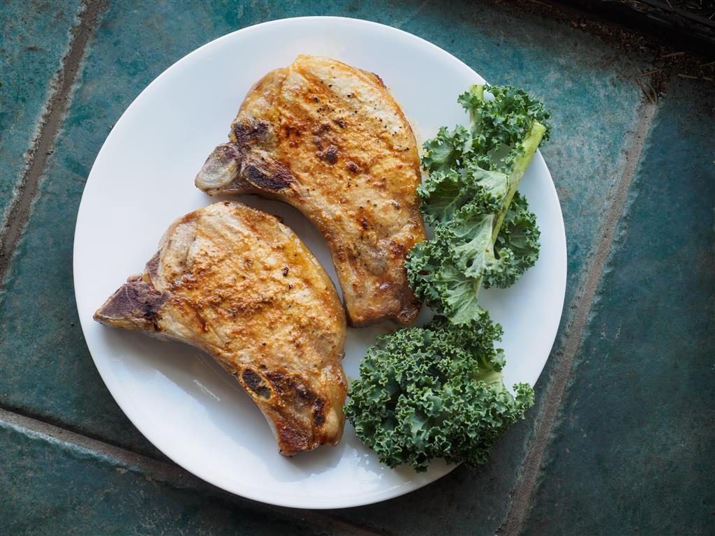 Two pork chops on a white plate with kale leaves, set on marble countertop